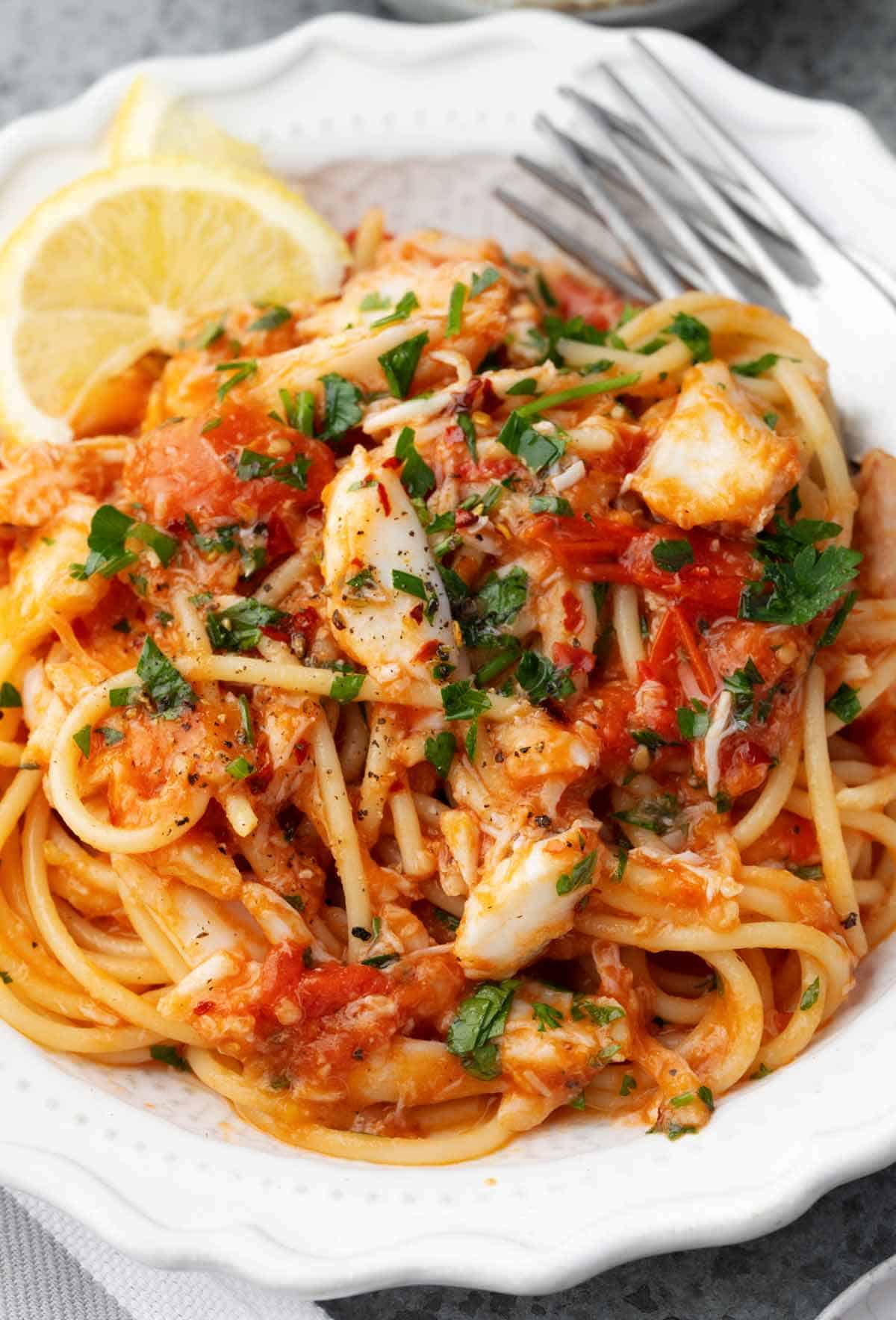 Close-up of crab pasta with spaghetti in a light tomato garlic sauce, topped with fresh parsley and served with lemon wedges on a white plate.