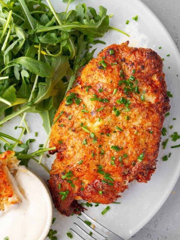 Crispy potato crusted ranch chicken breast topped with chopped chives, served with creamy ranch dipping sauce and fresh arugula on a white plate.