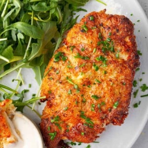 Crispy potato crusted ranch chicken breast topped with chopped chives, served with creamy ranch dipping sauce and fresh arugula on a white plate.