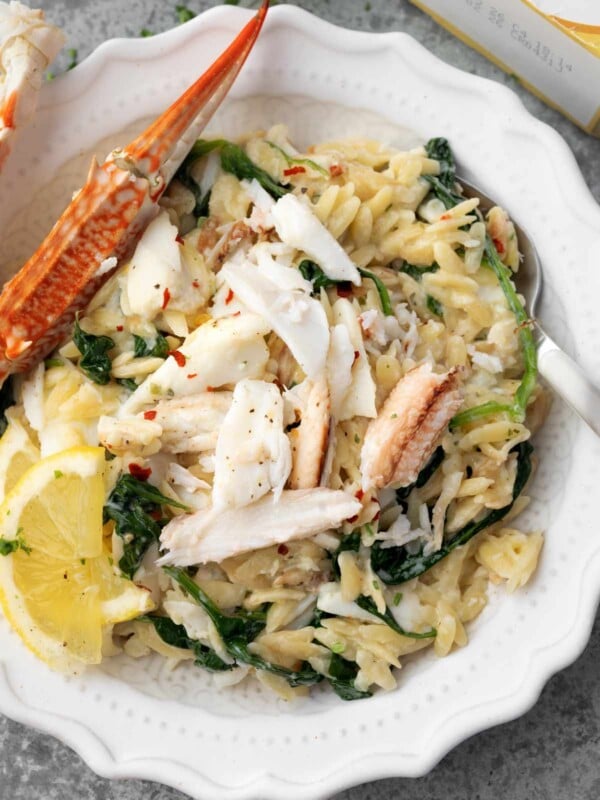 Creamy crab orzo pasta with spinach, lemon wedges, and red pepper flakes, topped with fresh crab meat and a crab claw on a white plate.