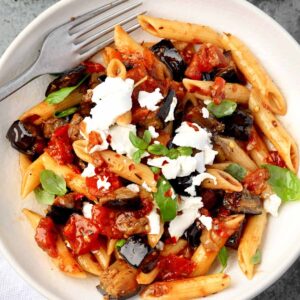 Pasta alla Norma with penne, roasted eggplant, tomato sauce, fresh basil, and crumbled ricotta salata served on a white plate.