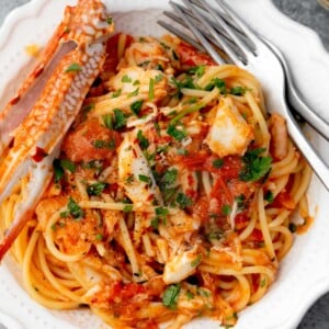 Close-up of spaghetti tossed in a light tomato garlic sauce with lump crab meat, fresh parsley, and red pepper flakes, served in a white bowl with lemon and crab legs on the side.