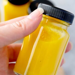 Close-up of a homemade ginger shot bottle held in hand, bright yellow citrus and turmeric wellness drink with fresh ginger.