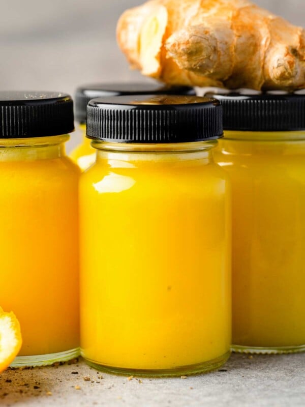 Fresh homemade ginger shots in small glass bottles with turmeric, citrus, and black lids, served with whole ginger root in the background.