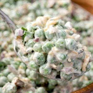 Close-up of creamy pea salad with bacon, cheddar, feta, and red onion on a spoon, coated in a tangy lemon dressing with fresh herbs.