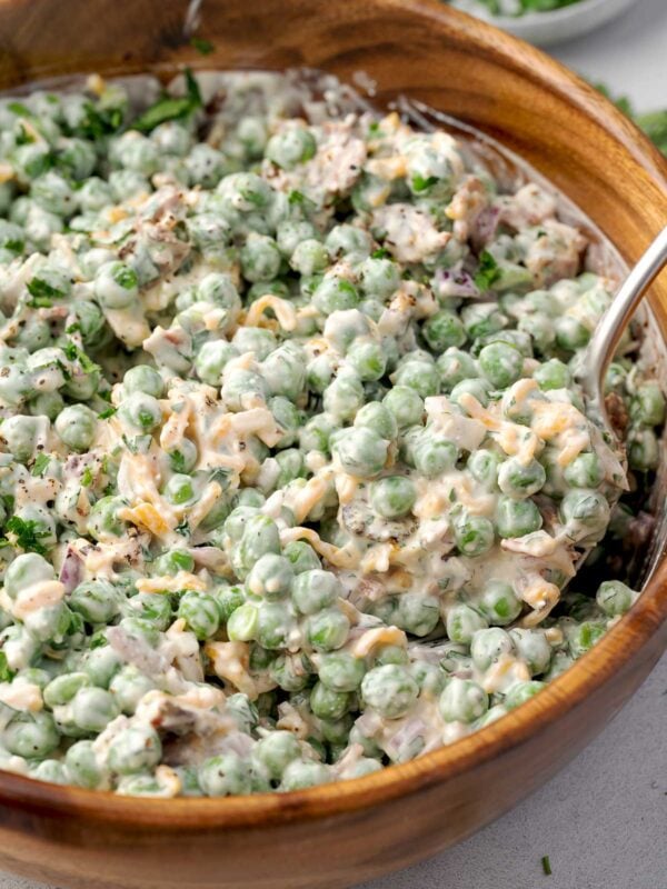 Creamy pea salad with bacon, cheddar, feta, and red onion in a wooden bowl, tossed in a tangy lemon dressing and garnished with fresh herbs.