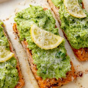 Baked salmon fillets topped with creamy broccoli pesto, fresh lemon slices, and grated Parmesan cheese on a sheet pan.