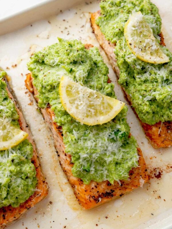 Baked salmon fillets topped with creamy broccoli pesto, fresh lemon slices, and grated Parmesan cheese on a sheet pan.