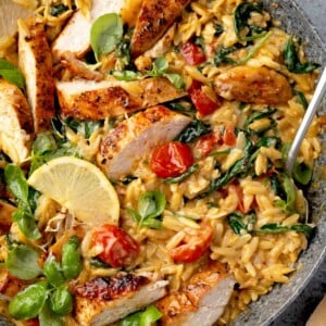 Creamy chicken and orzo skillet with sliced grilled chicken, cherry tomatoes, spinach, and basil in a rich garlic Parmesan cream sauce, garnished with lemon wedges.