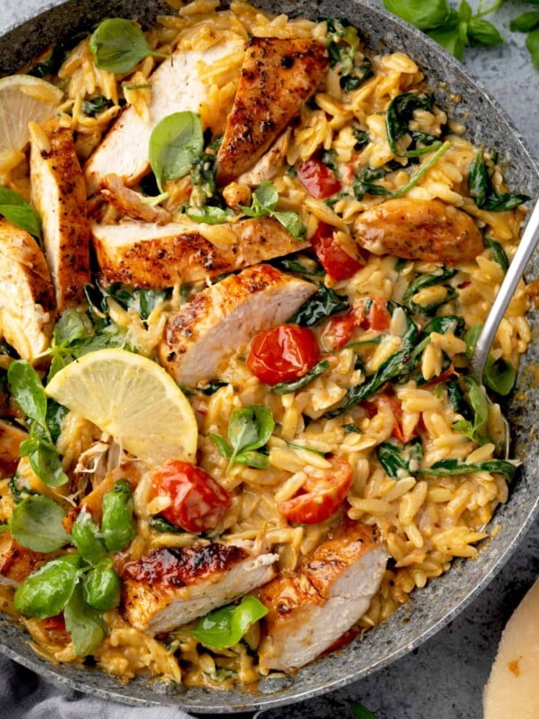 Creamy chicken and orzo skillet with sliced grilled chicken, cherry tomatoes, spinach, and basil in a rich garlic Parmesan cream sauce, garnished with lemon wedges.