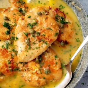 Golden seared chicken piccata in a skillet, topped with fresh parsley and capers, served in a rich lemon butter sauce with sliced lemon garnish.