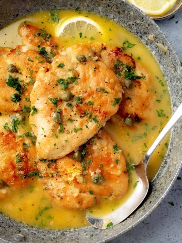 Golden seared chicken piccata in a skillet, topped with fresh parsley and capers, served in a rich lemon butter sauce with sliced lemon garnish.