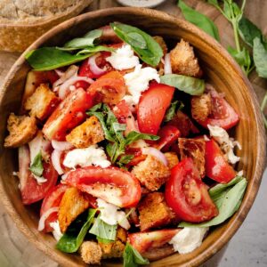 Panzanella salad with fresh tomatoes, crispy croutons, mozzarella, red onion, and basil in a wooden bowl, topped with olive oil and seasoning.