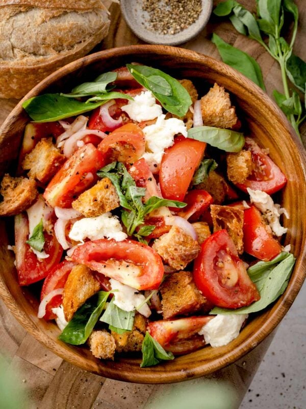 Panzanella salad with fresh tomatoes, crispy croutons, mozzarella, red onion, and basil in a wooden bowl, topped with olive oil and seasoning.