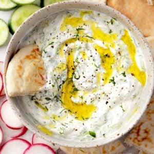 Creamy tzatziki sauce in a bowl with olive oil drizzle, fresh dill, cucumber slices, radishes, and warm pita bread on the side.