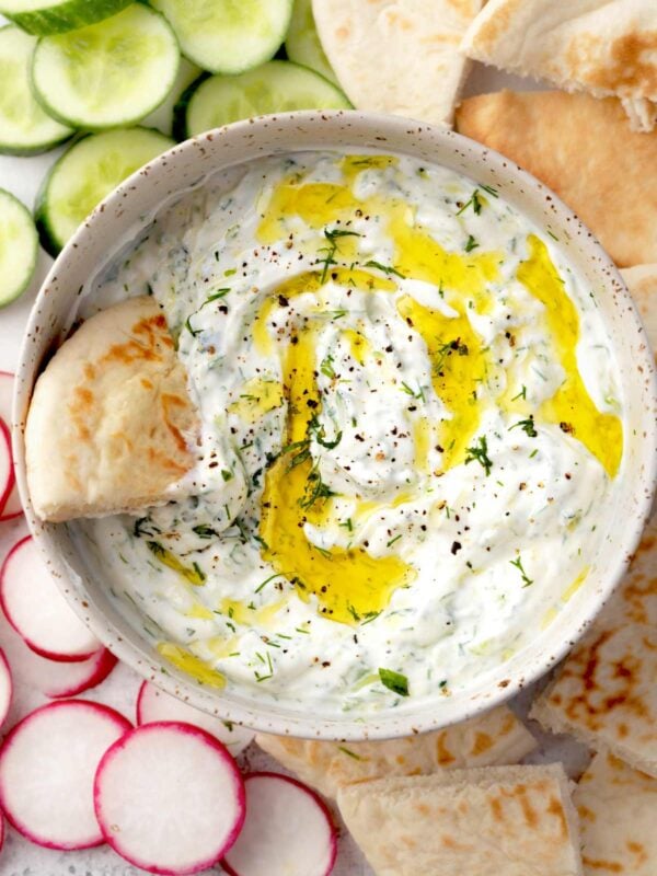 Creamy tzatziki sauce in a bowl with olive oil drizzle, fresh dill, cucumber slices, radishes, and warm pita bread on the side.