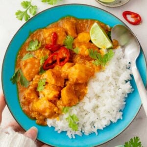 Thai coconut chicken curry served over jasmine rice with fresh cilantro, sliced red chili peppers, and lime wedges in a blue bowl.