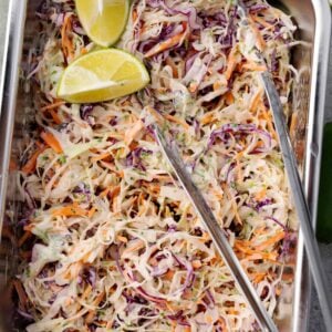 Mexican slaw made with shredded green and red cabbage, carrots, and a creamy lime-cilantro dressing, served in a metal tray with lime wedges on top.