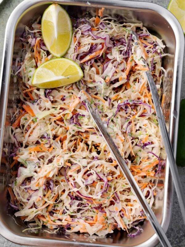 Mexican slaw made with shredded green and red cabbage, carrots, and a creamy lime-cilantro dressing, served in a metal tray with lime wedges on top.