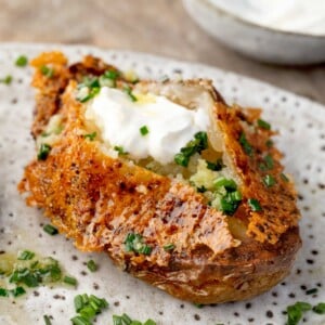 Close-up of a Parmesan crusted potato topped with sour cream, garlic-chive butter, and fresh chives, showcasing a crispy golden cheese crust and fluffy baked potato center.