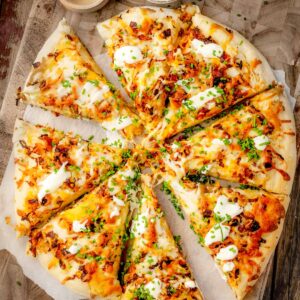 Pierogi pizza topped with creamy mashed potatoes, caramelized onions, crispy bacon, melted mozzarella and cheddar, sour cream, and fresh chives, sliced and served on a wooden pizza board.