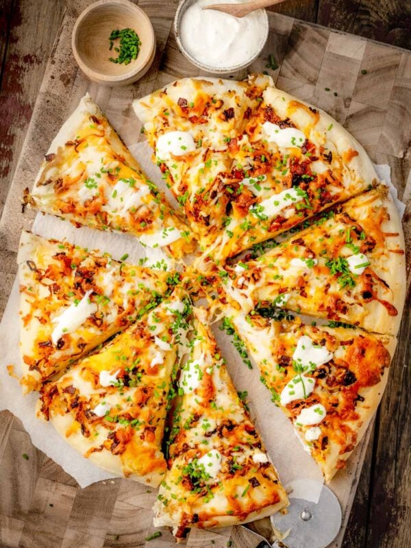 Pierogi pizza topped with creamy mashed potatoes, caramelized onions, crispy bacon, melted mozzarella and cheddar, sour cream, and fresh chives, sliced and served on a wooden pizza board.