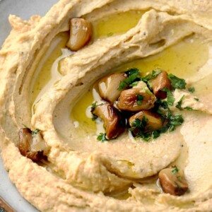Creamy roasted garlic hummus topped with caramelized roasted garlic cloves, fresh parsley, and extra virgin olive oil, served in a shallow bowl.