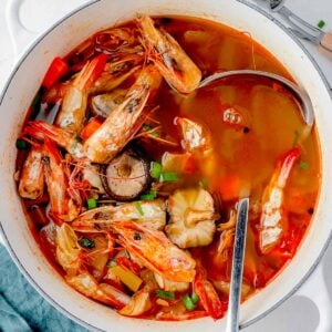 Homemade shrimp shell broth simmering in a Dutch oven with roasted shrimp heads and shells, mushrooms, garlic, celery, onions, and spices, with a ladle lifting the rich golden seafood stock.