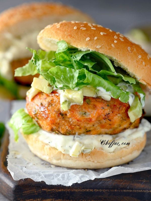 Salmon burger with tartar sauce, avocado, and crisp lettuce on a sesame seed bun.