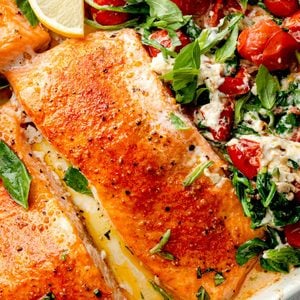 Close-up of baked Boursin salmon with roasted cherry tomatoes, creamy garlic and herb cheese sauce, wilted spinach, fresh basil, and lemon in a baking dish.