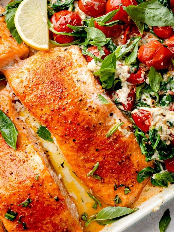 Close-up of baked Boursin salmon with roasted cherry tomatoes, creamy garlic and herb cheese sauce, wilted spinach, fresh basil, and lemon in a baking dish.