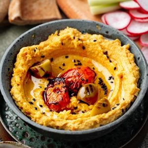 Close-up of roasted carrot hummus topped with caramelized carrots, roasted garlic, black sesame seeds, and olive oil, served with pita bread and fresh vegetables.