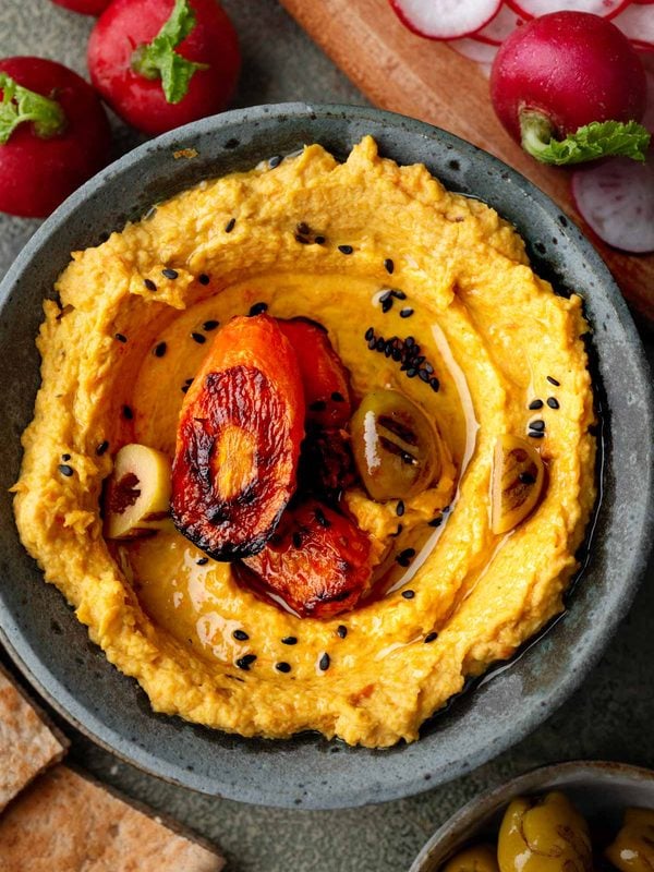 Overhead view of roasted carrot hummus topped with caramelized carrots, roasted garlic, black sesame seeds, and olive oil, served with pita bread, radishes, and olives.